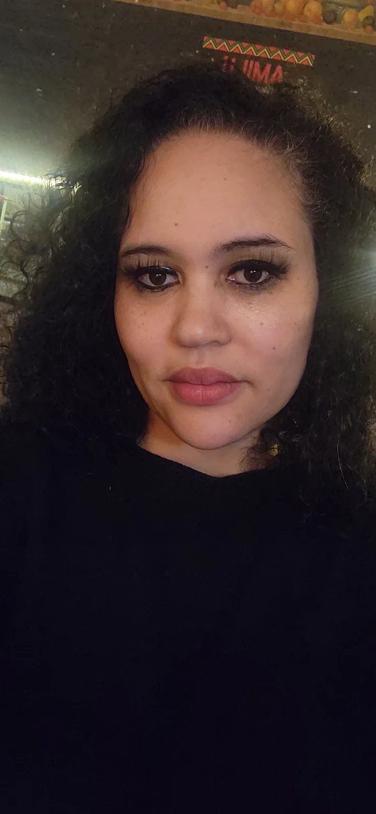 A close-up selfie of a woman with dark curly hair, wearing heavy black eyeliner and a neutral lip color, looking directly at…