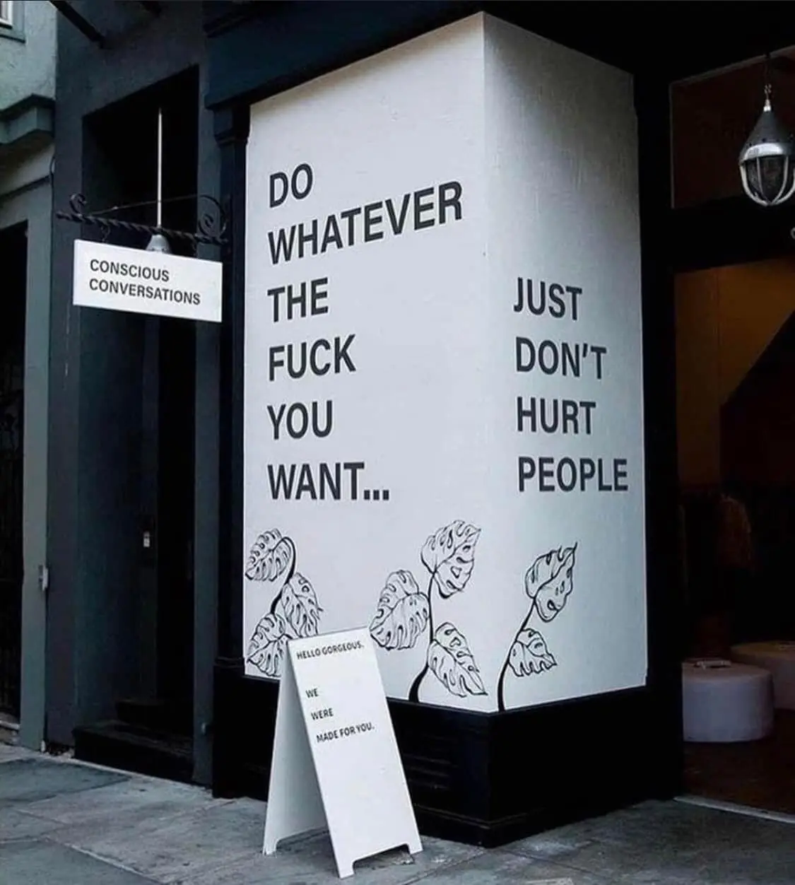 A white-painted corner of a building features bold black text reading 'DO WHATEVER THE FUCK YOU WANT... JUST DON'T HURT…