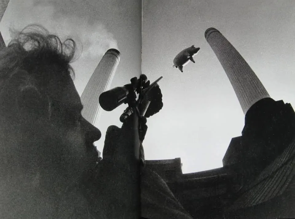 A black-and-white photo shows a person in profile holding a rifle with a scope, aiming upward at a pig flying between two…