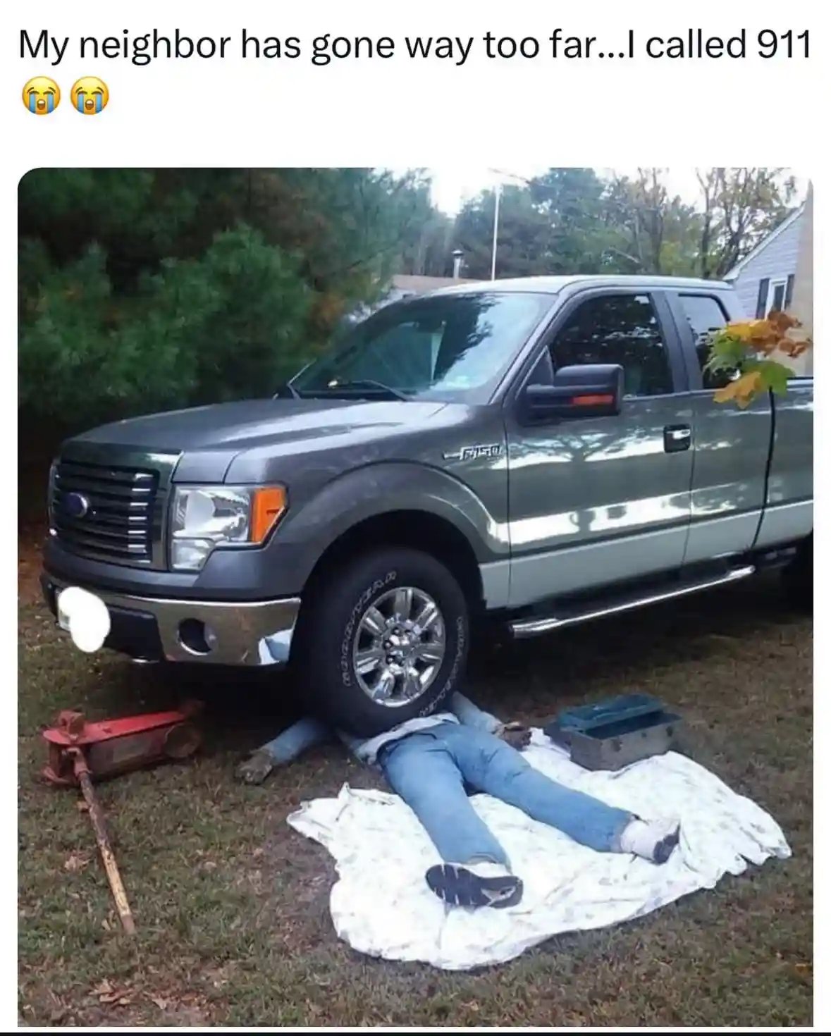 A person lies motionless on a white cloth on grass beneath a gray Ford F-150 pickup truck, with a red floor jack nearby. The…