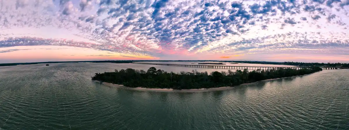 A panoramic aerial photo of a serene waterway at sunset, featuring a long bridge stretching across the horizon, a small…