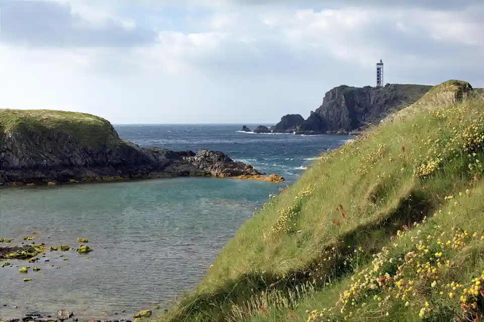 A scenic coastal landscape featuring a lighthouse on a distant rocky cliff, calm turquoise water in the foreground, and a…