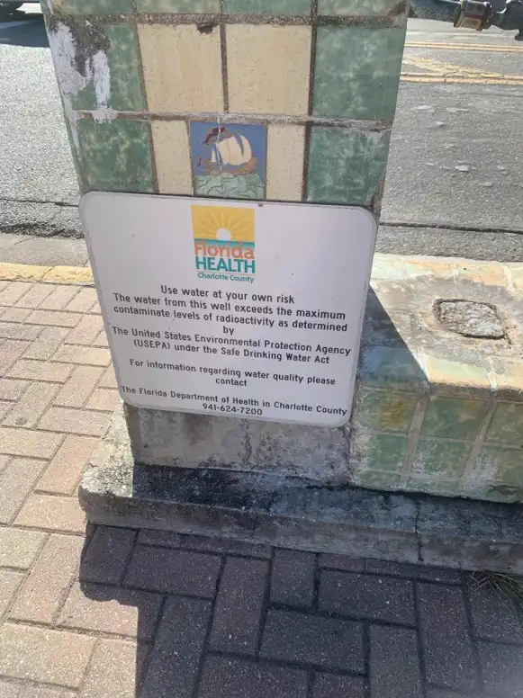 A white sign from Florida Health Charlotte County is mounted on a tiled well structure. The sign warns that the well water…