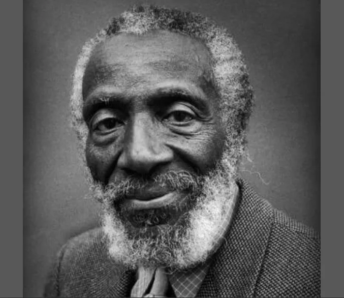 A black-and-white portrait of an elderly Black man with a white beard and mustache, wearing a suit and tie, smiling gently…