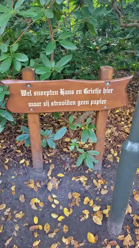 A wooden sign in a park surrounded by green bushes and fallen autumn leaves reads 'Wel snoepten Hans en Grietje hier maar…
