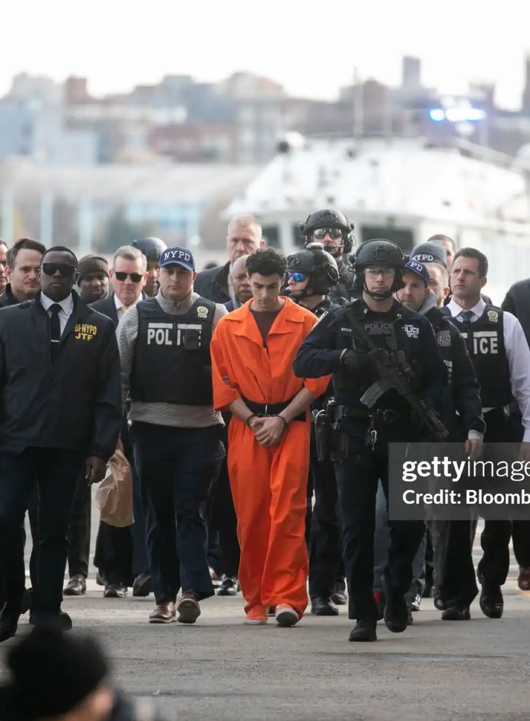 A young man in an orange prison jumpsuit is being escorted by numerous heavily armed police officers, including NYPD and FBI…