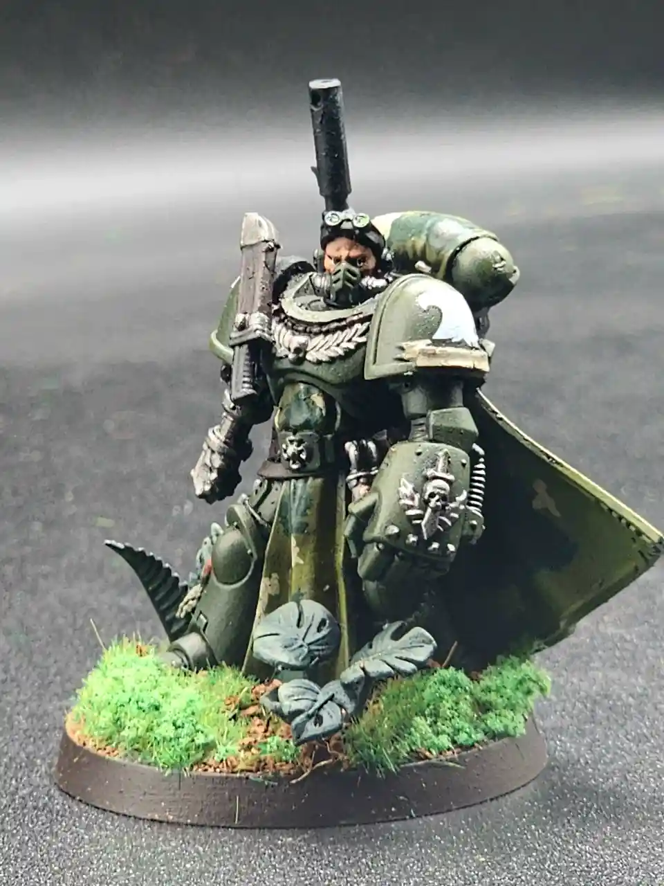 A heavily armored, green-painted miniature warrior from Warhammer 40,000 stands on a circular base with green grass and…