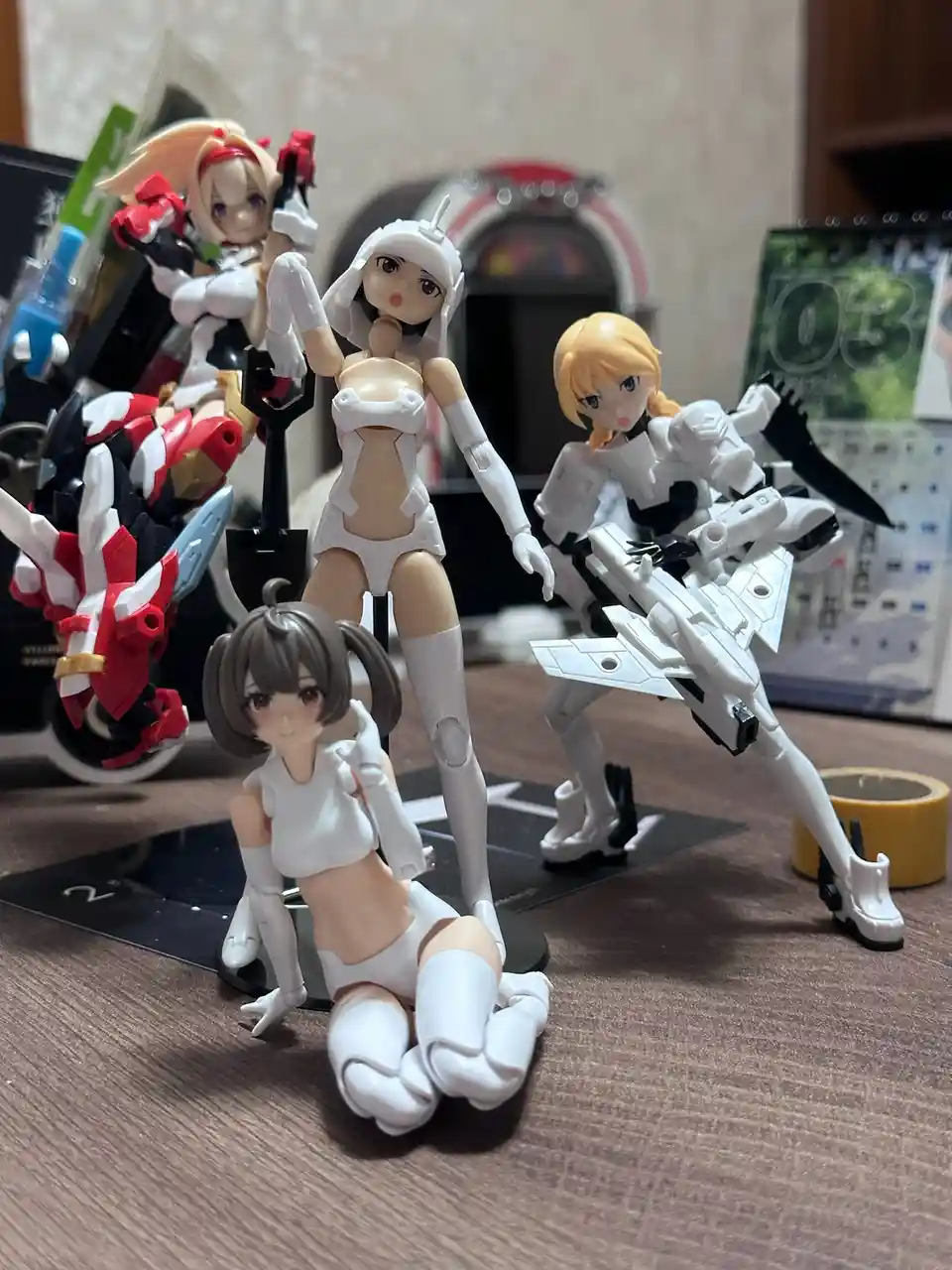 Four anime-style action figures in white outfits are posed on a wooden desk, with one kneeling in the foreground and three…