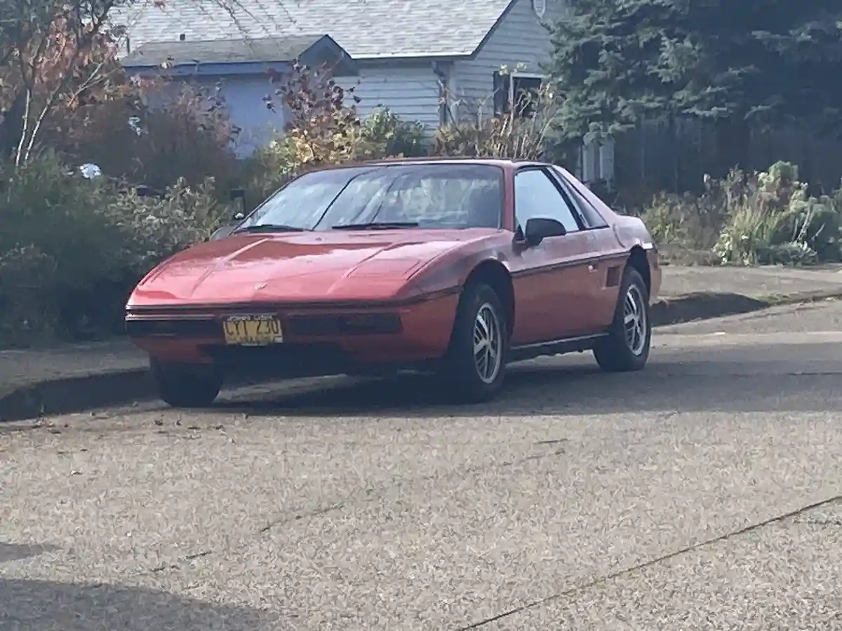 A red, vintage sports car, likely a Mazda RX-7, is parked on a residential street with a house and trees in the background…