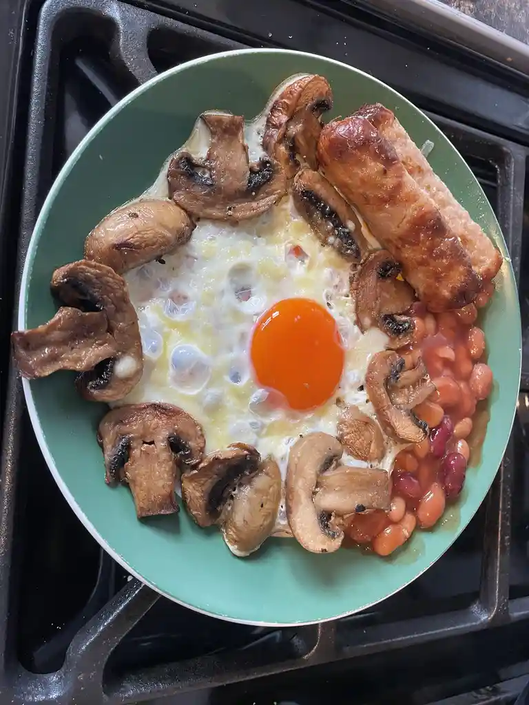 A green plate on a stovetop holds a fried egg with a bright orange yolk at the center, surrounded by sautéed mushrooms, two…