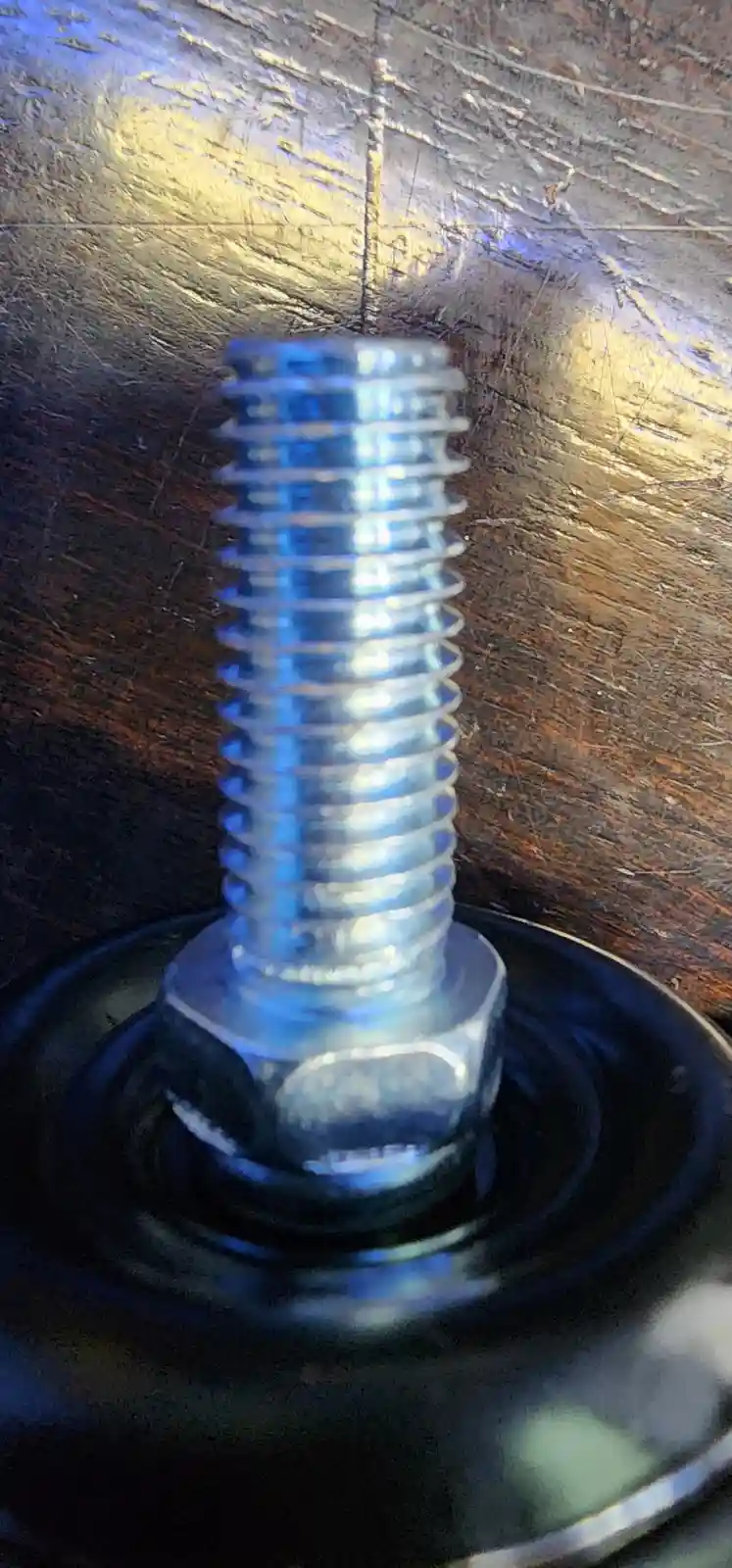 A close-up photo shows a shiny metal bolt with a hexagonal head, standing upright in a black circular washer on a scratched…