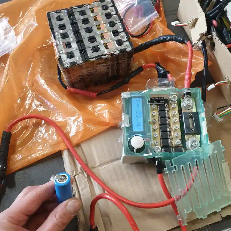 A hand holds a small blue cylindrical battery while red wires connect it to a custom-built electronic device with a digital…