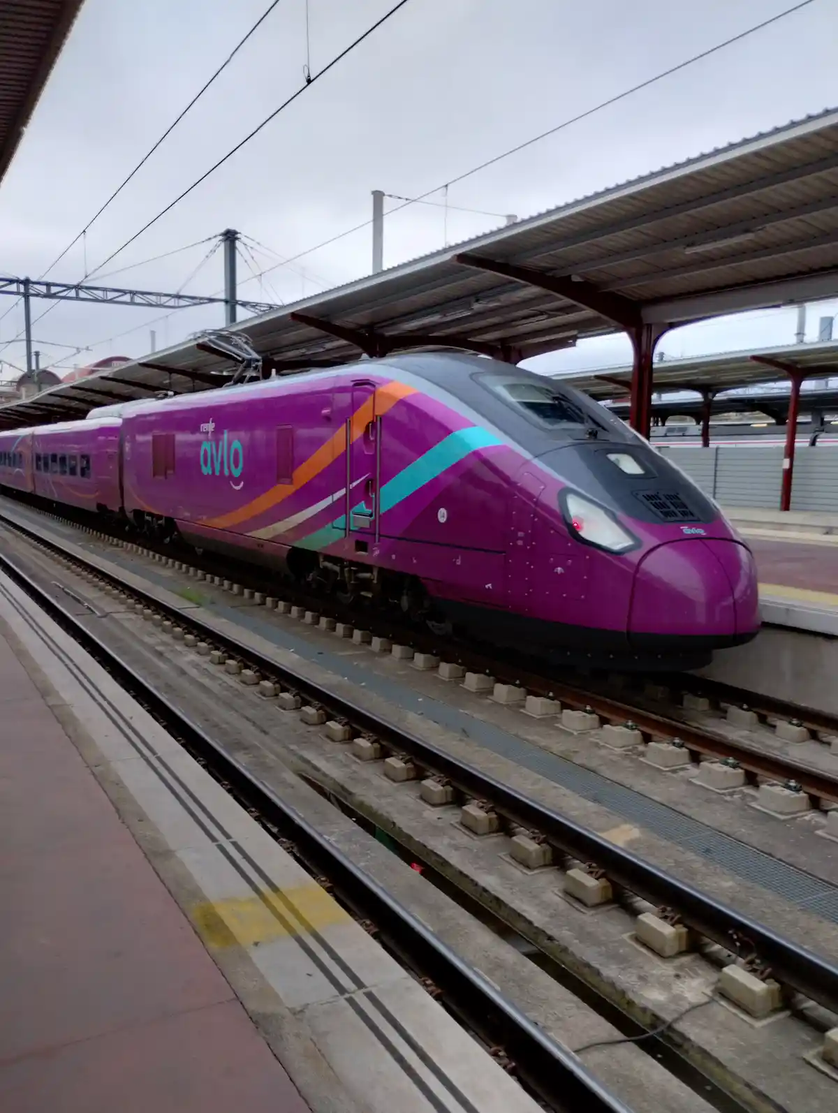 A purple and teal Renfe Avlo train is stationary at a station platform under an overcast sky. The train's modern design…