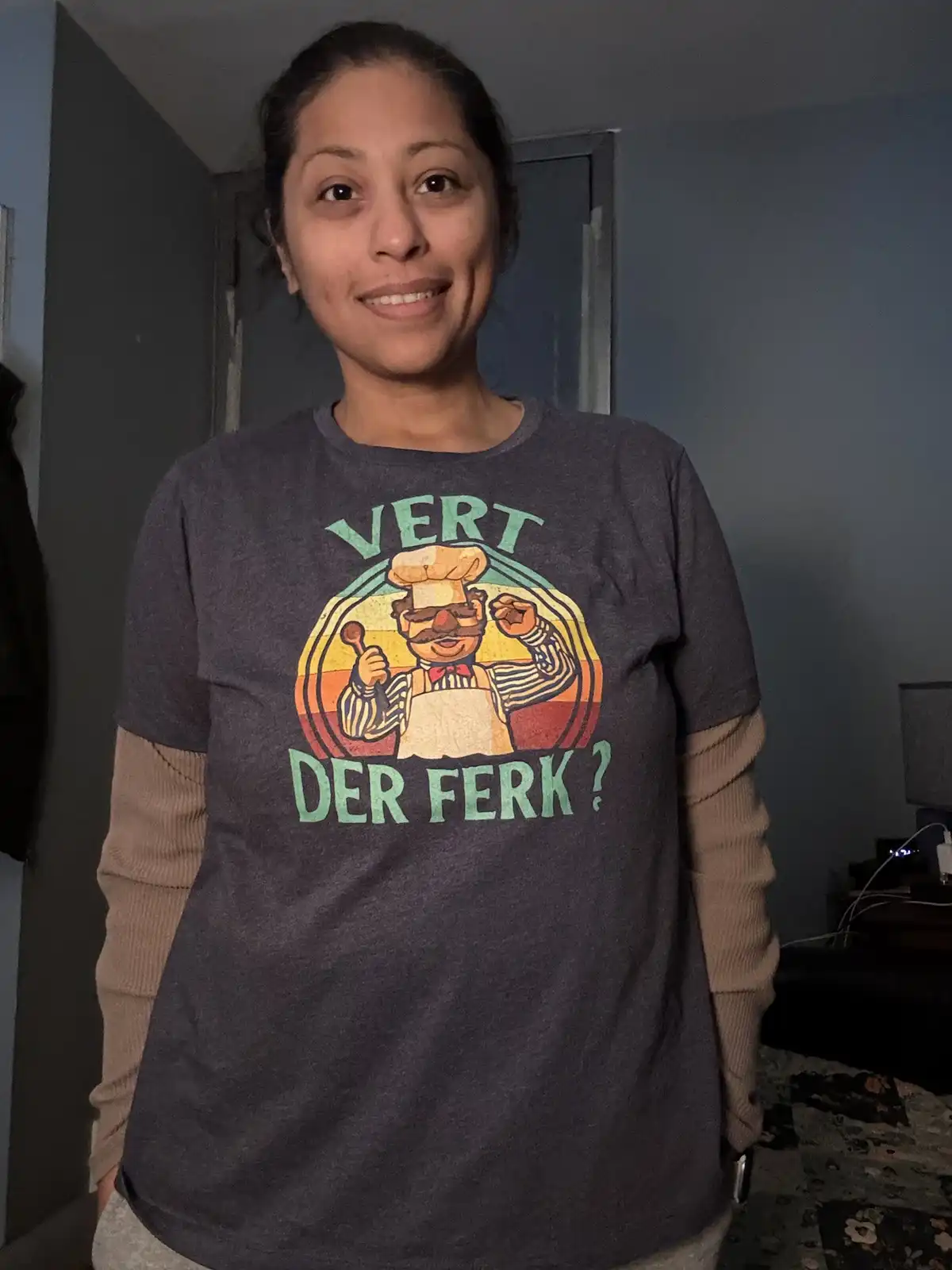 A woman smiles while wearing a dark gray t-shirt featuring a vintage-style graphic of a muscular chef with a mustache…