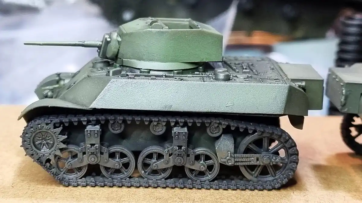 A close-up photograph of a detailed green military tank model with visible treads and wheels, sitting on a wooden surface…