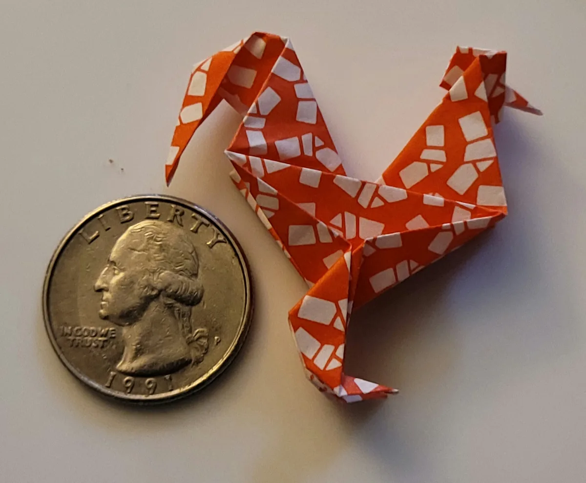A small orange and white patterned origami rooster sits next to a 1991 U.S. quarter on a plain surface, with the text 'i…