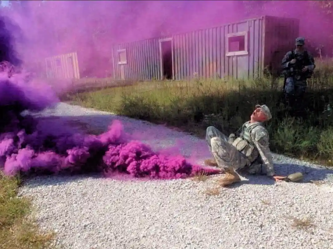 A soldier in camouflage uniform sits on a gravel path, looking up at a large cloud of vibrant purple smoke billowing from a…