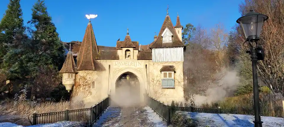 A picturesque, snow-dusted European-style castle gate with ornate turrets and a stone archway, partially obscured by mist…