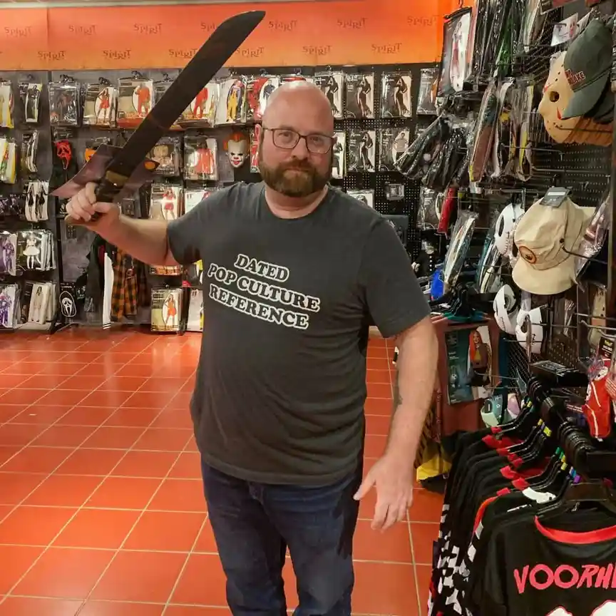 A bald man with glasses and a beard stands in a Spirit Halloween store, holding a large prop machete. He wears a dark gray…