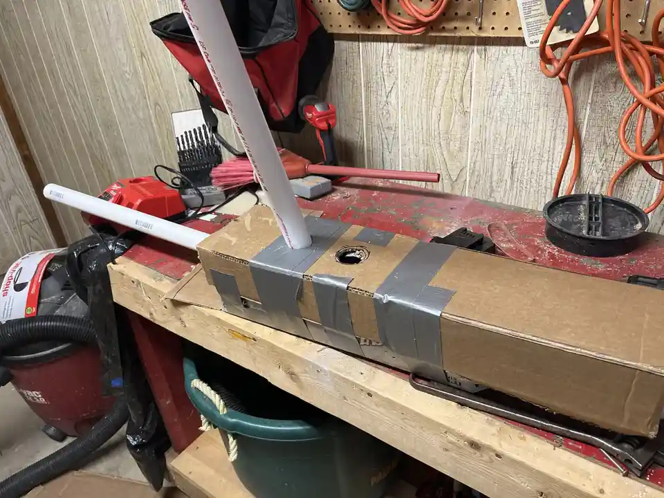 A cardboard box taped with gray duct tape sits on a cluttered red workbench in a garage, with white PVC pipes inserted into…