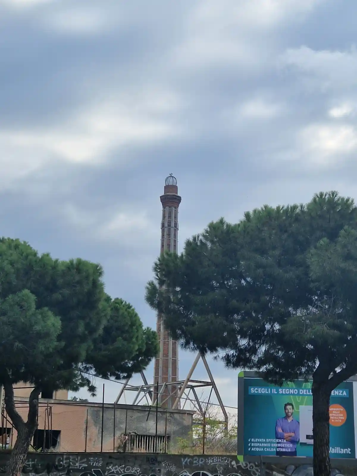 A tall, brick industrial chimney with a metal lattice support structure rises behind two large, leafy trees under a cloudy…