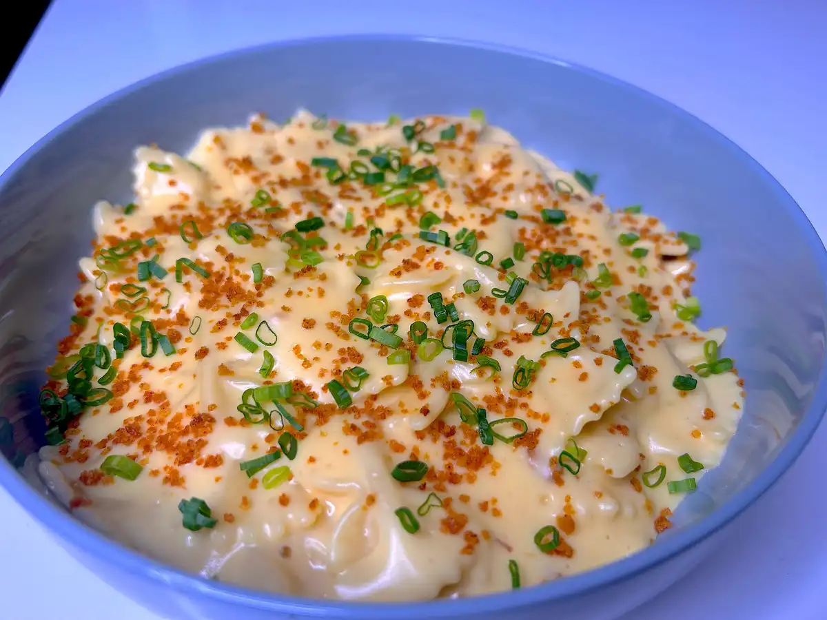 A close-up photo shows a light blue bowl filled with creamy macaroni and cheese, topped with golden-brown breadcrumbs and…