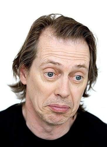 A man with messy brown hair and a black shirt stares with wide, startled eyes and a slightly downturned mouth, conveying…