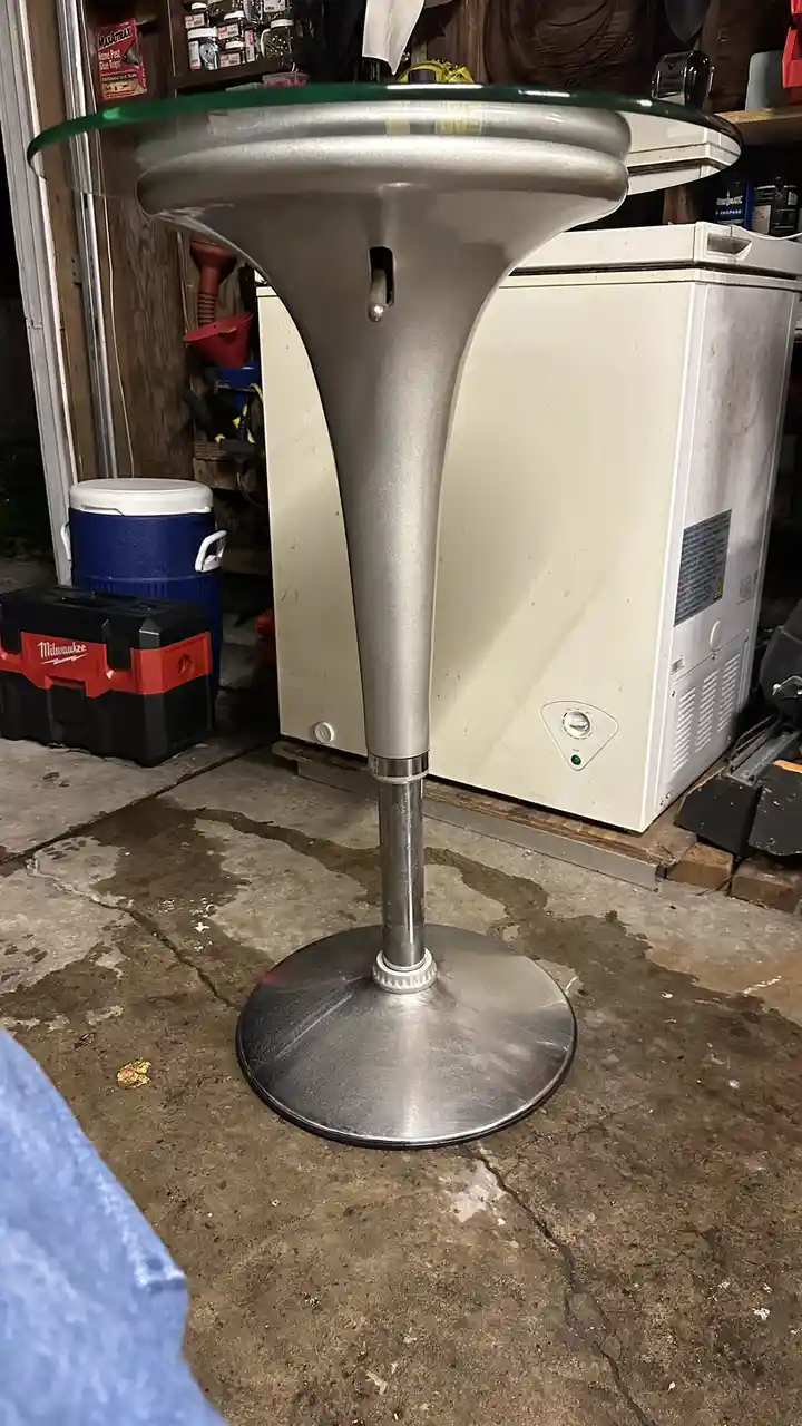 A silver, funnel-shaped table with a glass top sits in a cluttered garage next to a white freezer and a blue cooler. The…