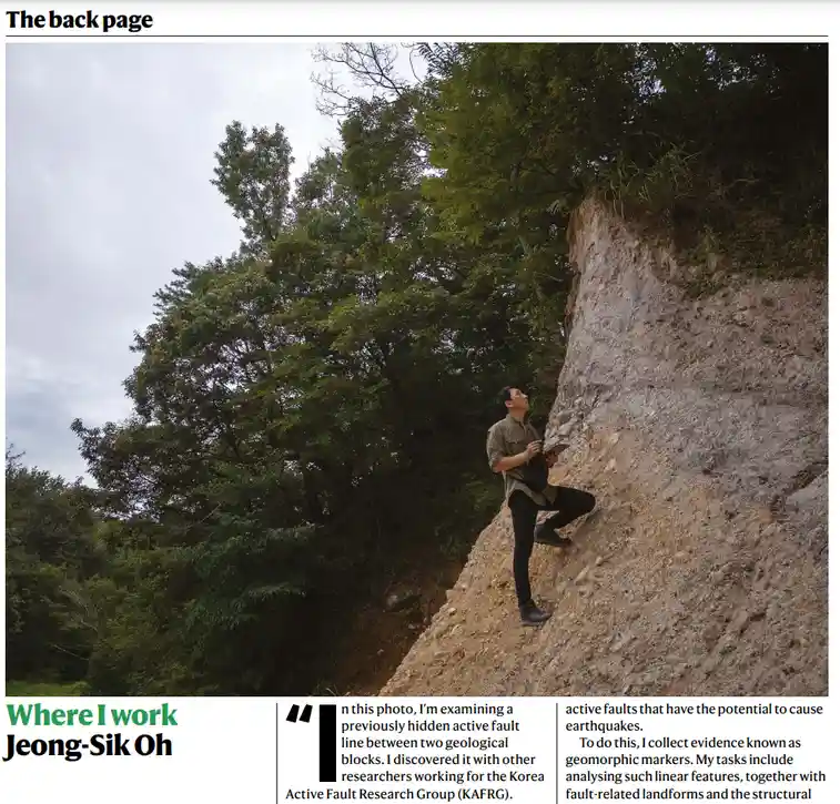 A man in a beige shirt and black pants is climbing a steep, exposed rock face in a forested area, looking upward. The image…