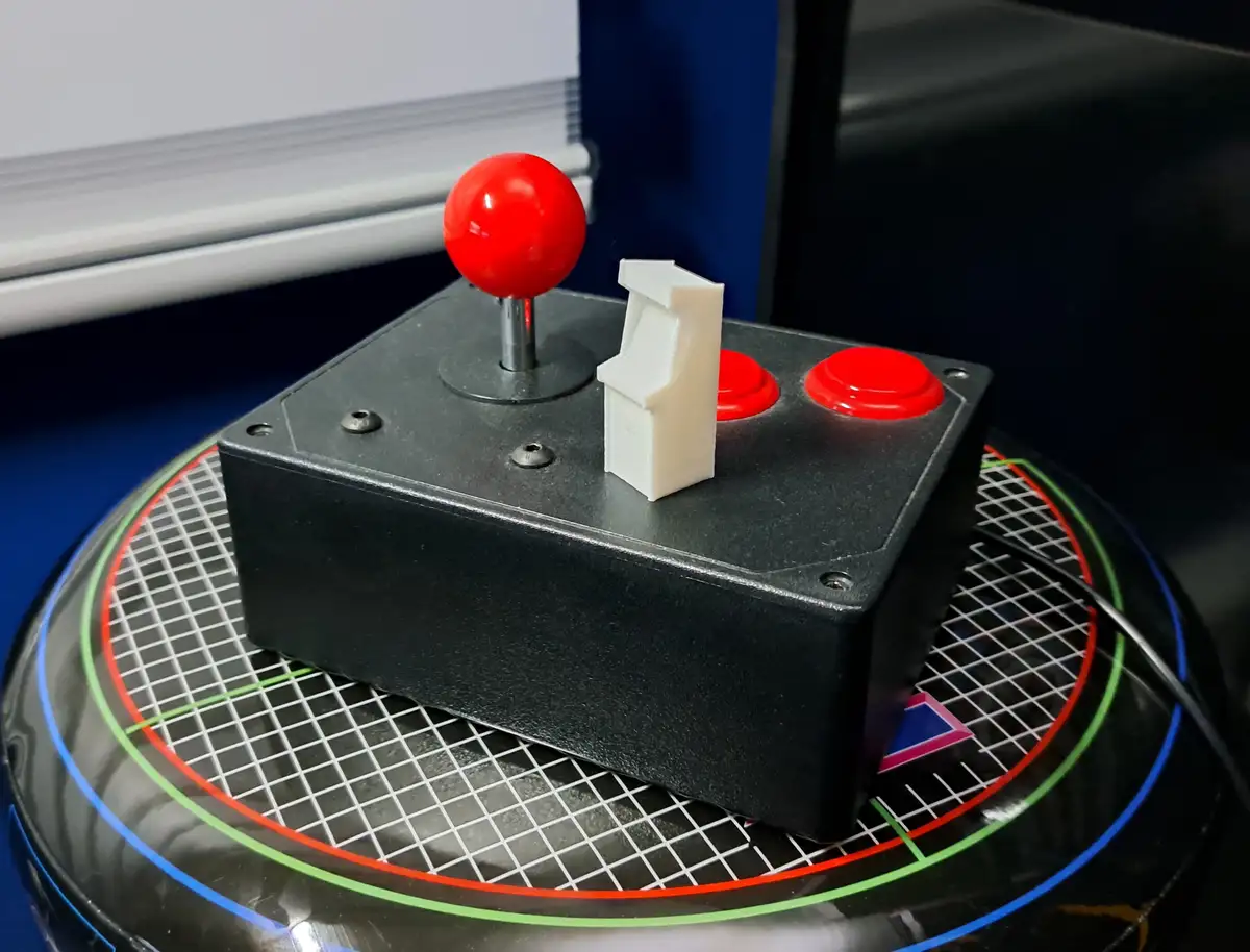 A close-up photo shows a black arcade-style game controller with a red joystick and two red buttons, resting on a circular…