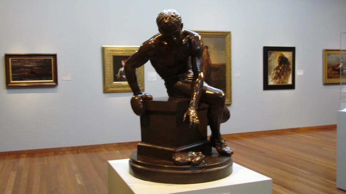 A bronze statue of a muscular boxer sitting on a pedestal in an art gallery, wearing boxing gloves and shoes, with several…