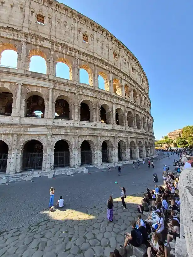 A wide-angle photo shows the massive, multi-tiered Colosseum in Rome under a clear blue sky, with tourists scattered around…