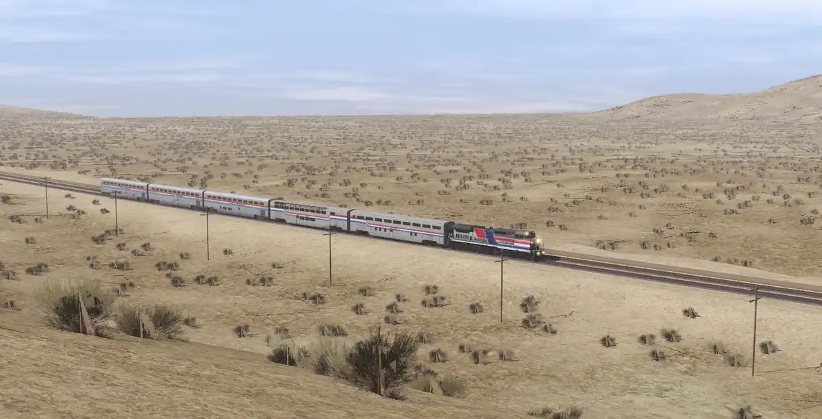 A long Amtrak train travels through a vast, arid desert landscape with sparse vegetation and rolling hills under a pale blue…