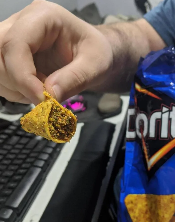 A hand holds a partially eaten, crumpled Dorito chip that appears to have been filled with a dark, textured substance…