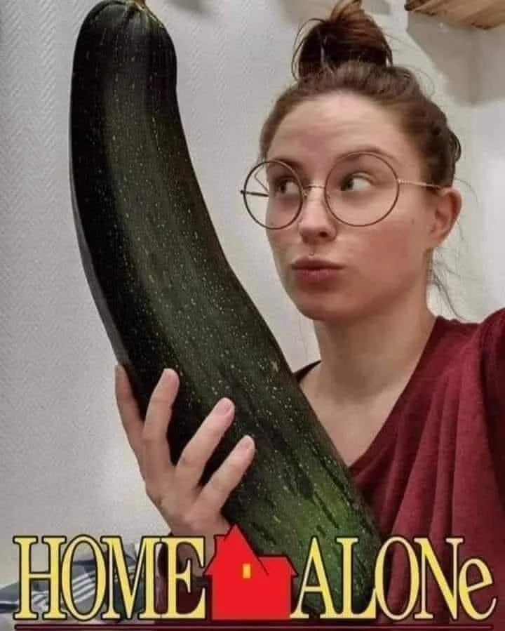 A woman with glasses and her hair in a bun holds an extremely large, dark green zucchini. She looks sideways with a…