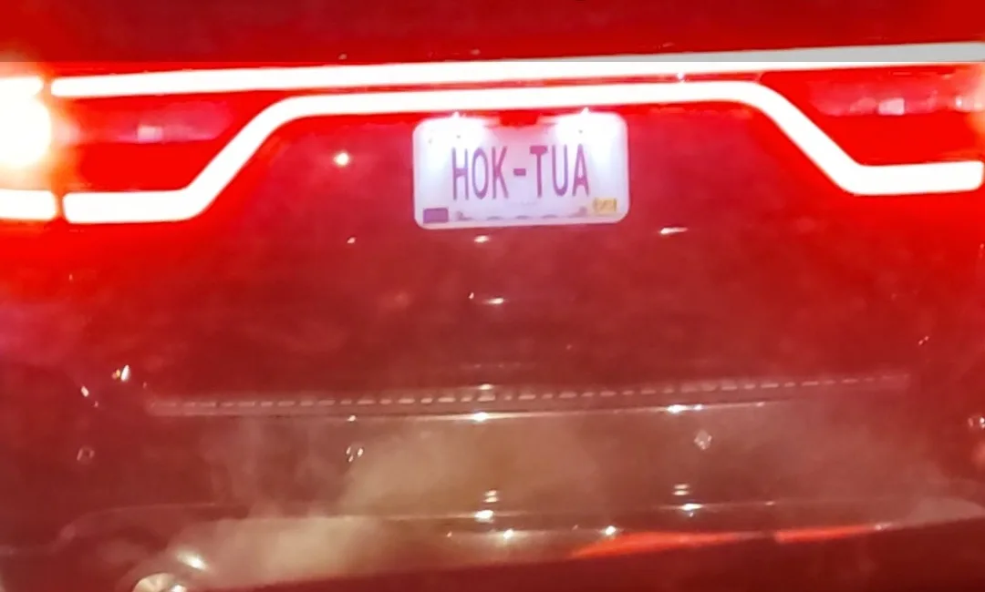 A close-up of the rear of a red car at night, showing its illuminated taillights and a license plate that reads 'HOK-TUA'…