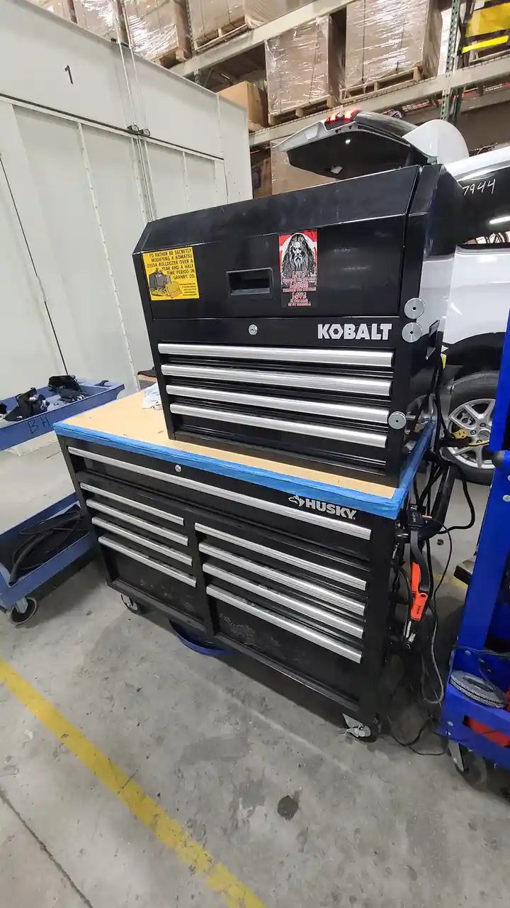 A black Kobalt tool chest sits atop a Husky tool cart in a warehouse, with a yellow sticker on the chest reading 'I'D RATHER…