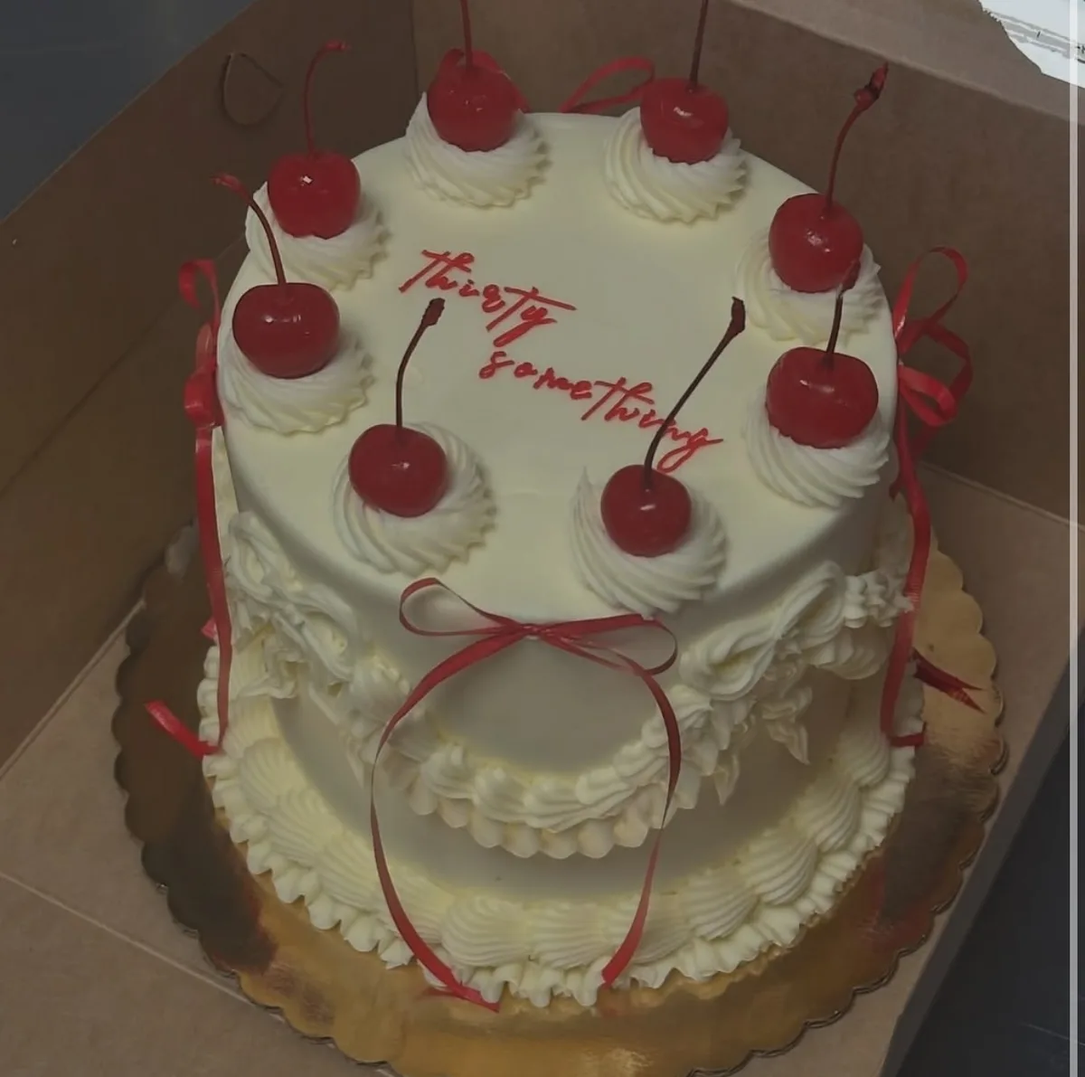 A two-tiered white cake with white frosting, red cherries on top of whipped cream swirls, and red ribbons tied around it…