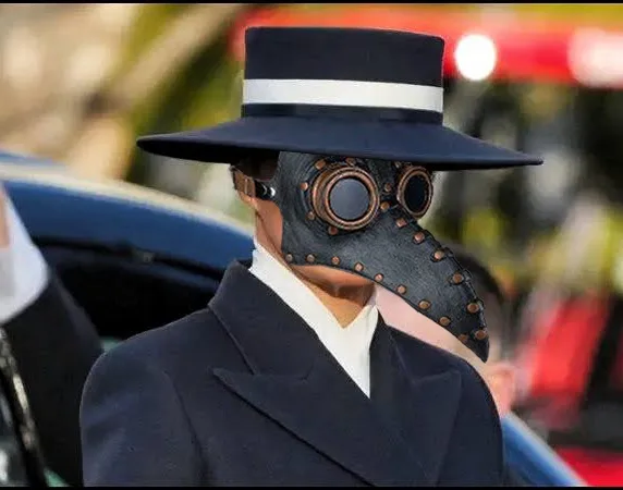 A person wearing a dark blue suit and white shirt is shown from the chest up, wearing a black plague doctor mask with a long…