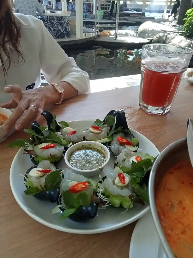 A person sits at a wooden table in a restaurant with a pond in the background, in front of them is a plate of fresh shrimp…