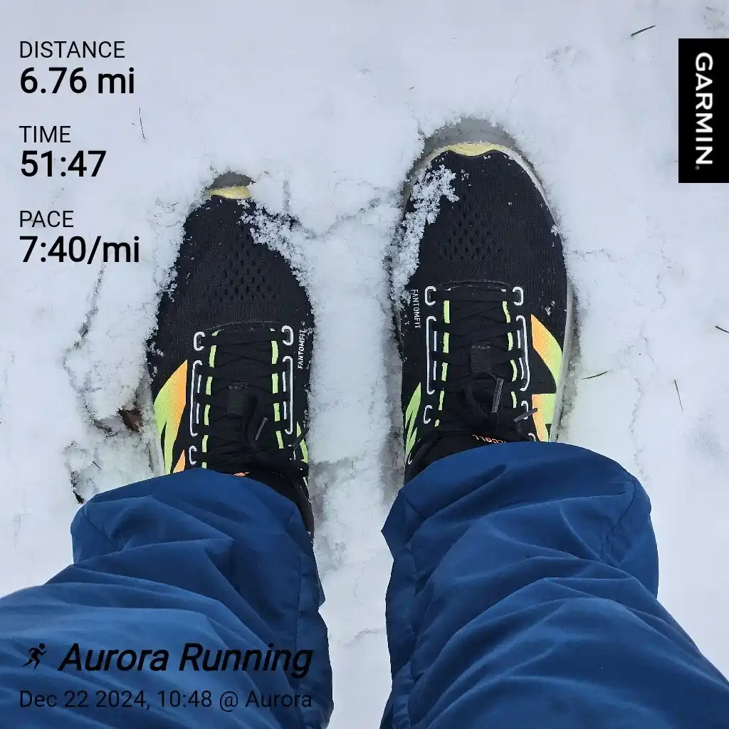 A first-person view of someone's feet in black and neon green running shoes standing in snow, with a Garmin fitness tracker…