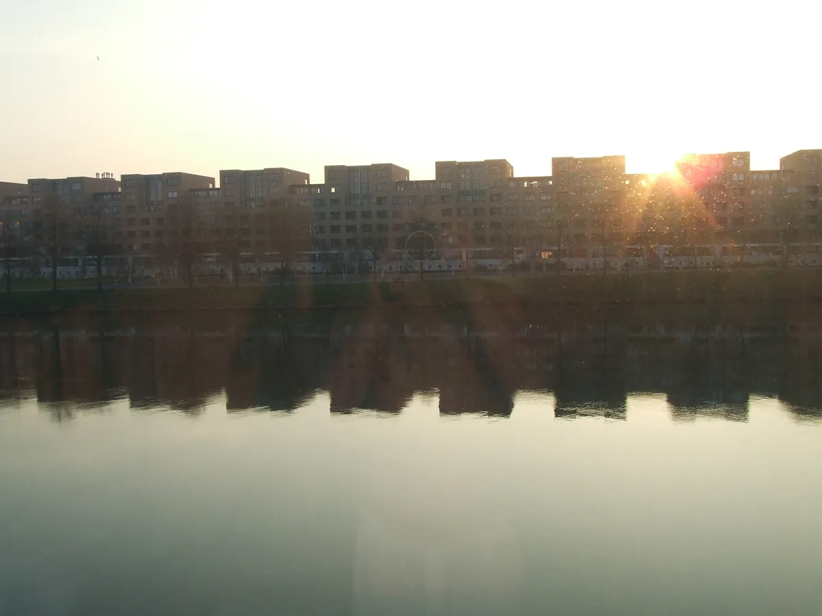 A serene sunset photo shows modern apartment buildings reflected in calm water, with the sun creating lens flare. Overlaid…
