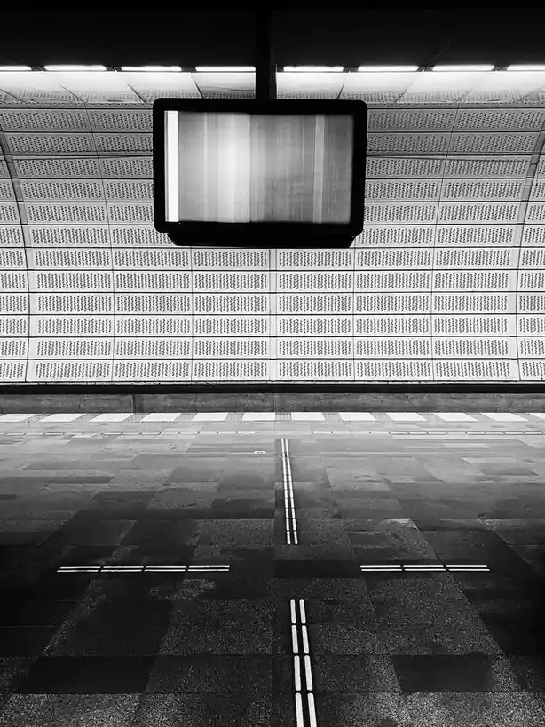 A black-and-white photo of an empty, modern subway or train station platform with a large, blank digital display screen…