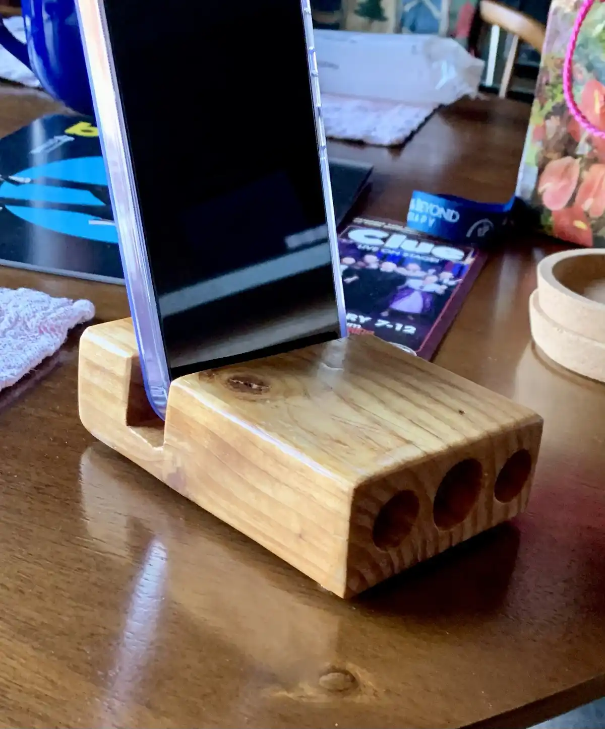 A smartphone in a clear case rests in a wooden stand that is shaped like a six-sided die, with three circular holes visible…