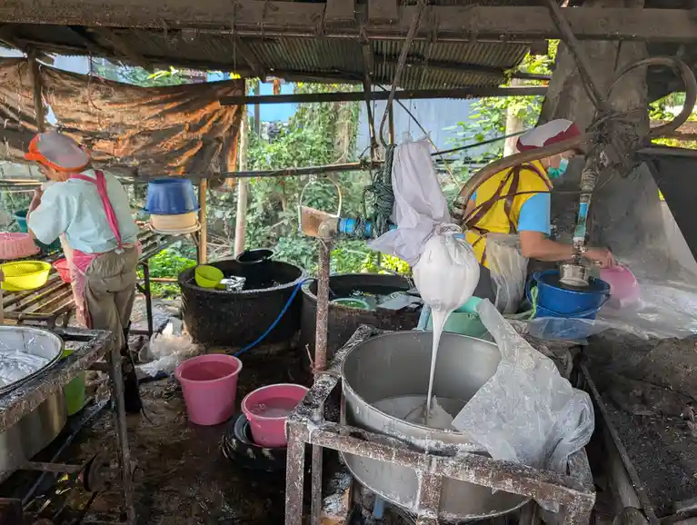 two workers in a rustic, wet outdoor kitchen pour thick white liquid from a bag into a metal tub, surrounded by buckets and…