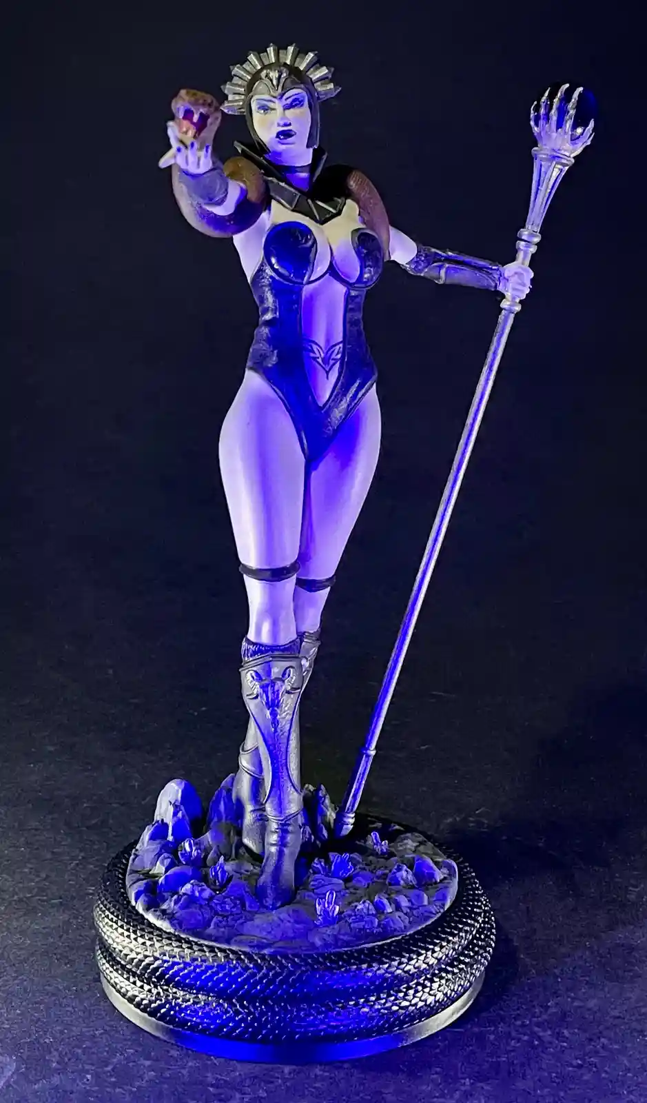 A statue of a female superhero or villain in a revealing blue and silver costume, holding a staff and a glowing orb…
