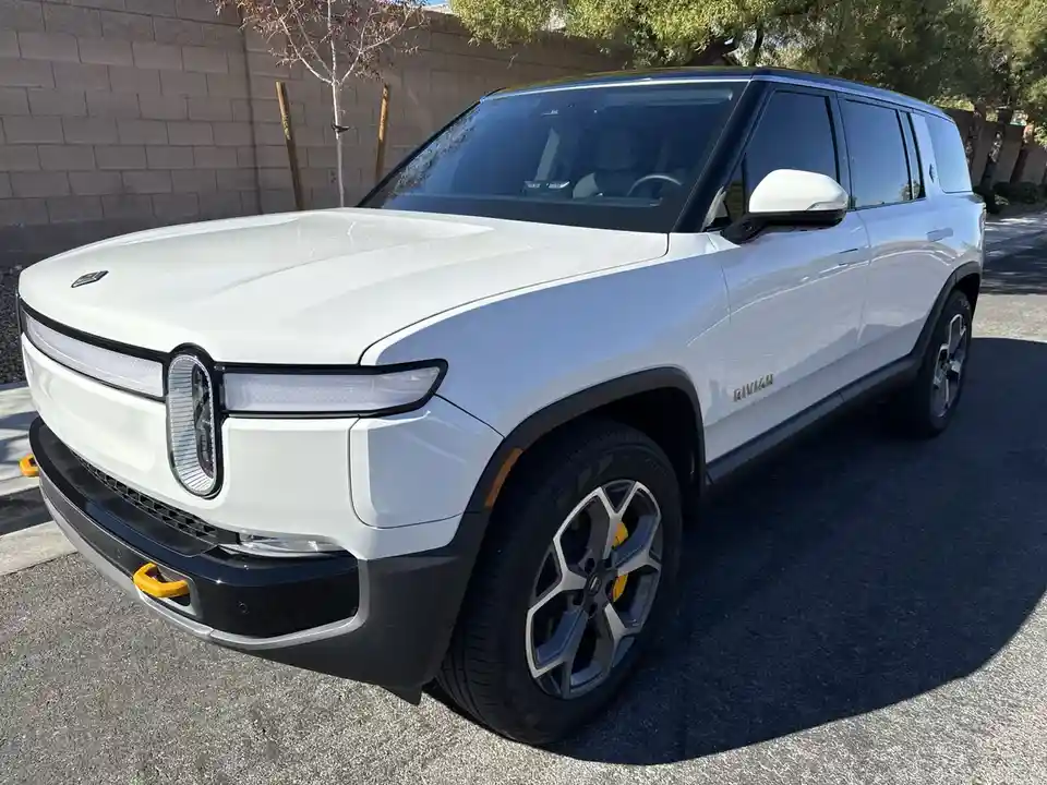 A white Rivian R1T electric pickup truck is parked outdoors on asphalt, angled to show its front and side. The vehicle…
