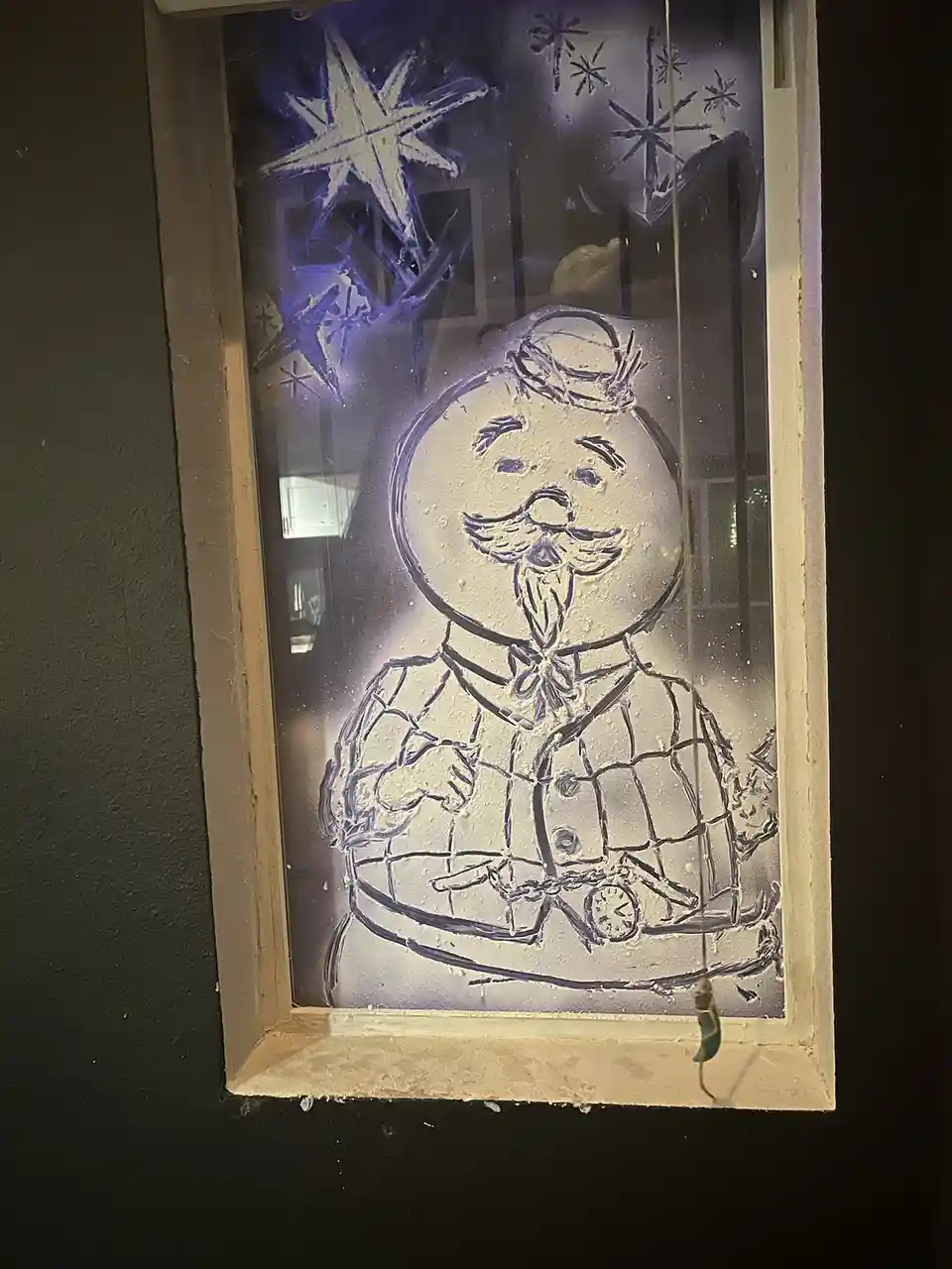 A frosty window features a hand-drawn snowman with a mustache, bow tie, and checkered coat, surrounded by snowflake…