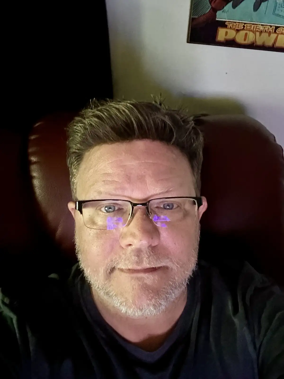 A man with glasses and a beard sits in a dark room, looking directly at the camera with a weary expression. Purple light…