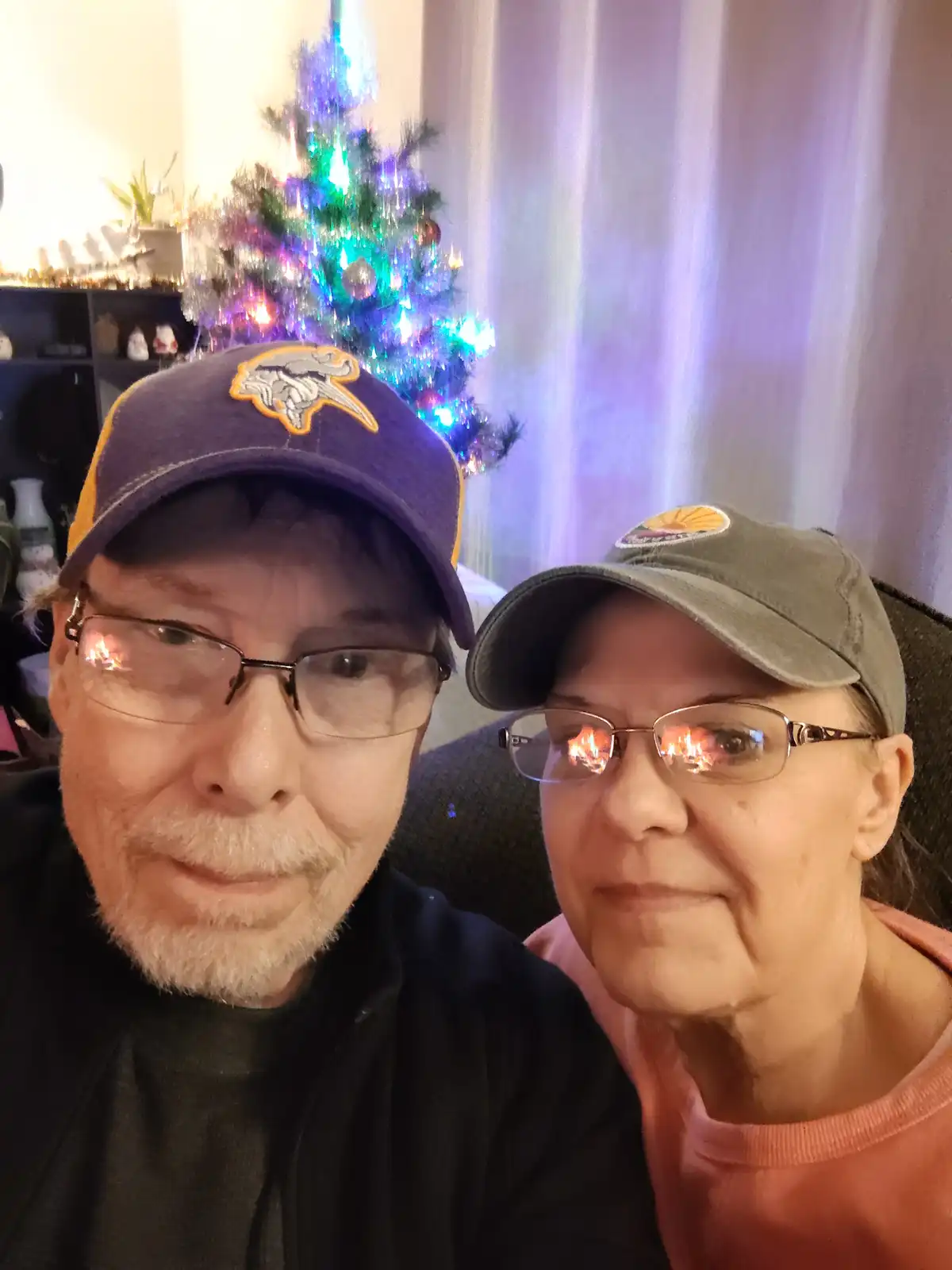An older couple poses for a selfie in front of a lit Christmas tree; the man wears a purple Minnesota Vikings baseball cap…